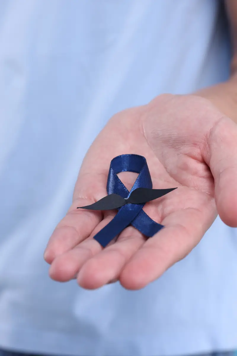 Prostate cancer awareness. Man holding blue ribbon with fake mustache on color background, closeup Prostate cancer awareness. Man holding blue ribbon with fake mustache on color background, closeup